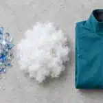 Recycled polyester fabric flat-lay: clear PET bottle flakes, white polyester fibre and a folded teal softshell jacket with a recycled-content hangtag